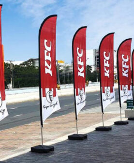 Røde beachflagg med KFC-logo på rekke langs et fortau for maksimal synlighet ved vei.
