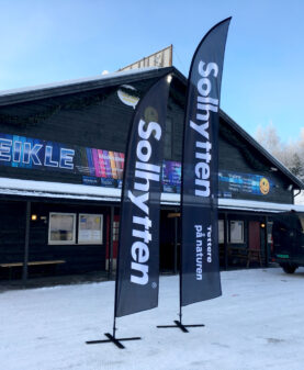 Grå beachflagg med Solhytten-logo montert utenfor bedriftslokale i vinterlandskap.