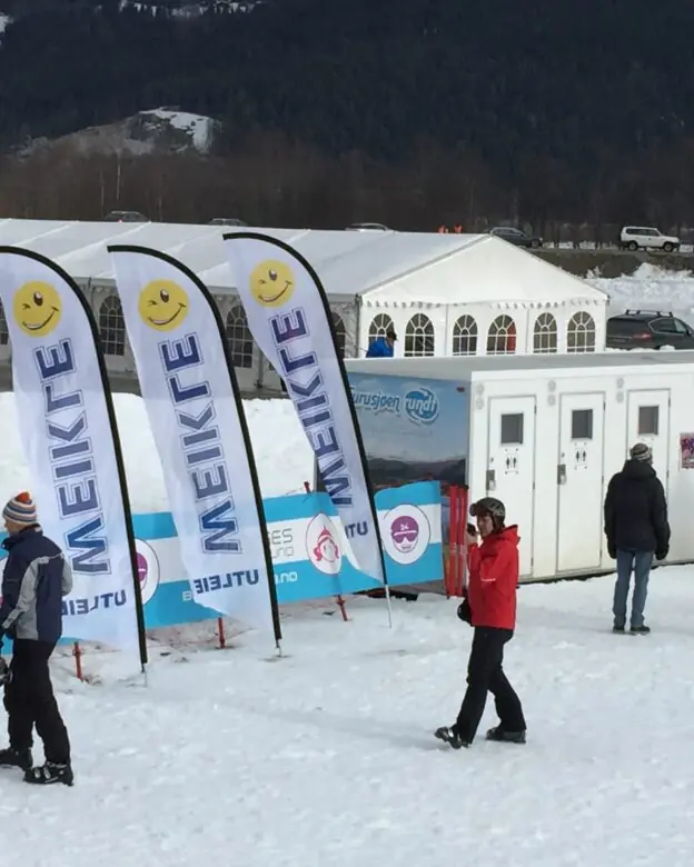 Hvite beachflagg med logo og smilefjes plassert foran et stort arrangementstelt i fjellet.