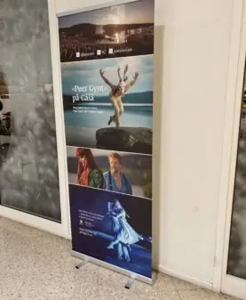En rollup-banner står på gulvet og viser reklame for Peer Gynt på Gålå med flere bilder fra forestillingen.