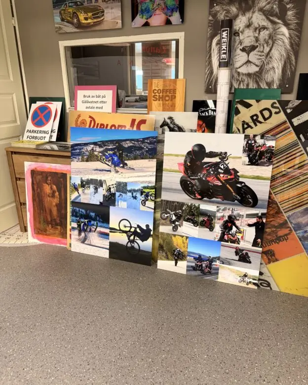 Flere store bilder og plakater med motiver av motorsykler og ekstremsport står lent mot en vegg i et rom.