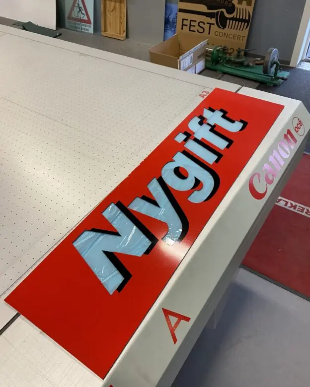 Et stort, rødt klistremerke med ordet "Nygift" i lyseblå skrift ligger på et arbeidsbord.