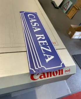 Flere blå klistremerker med hvit tekst "CASA REZA" ligger stablet på en Canon Océ plotter.