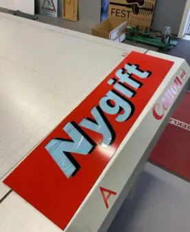 Et stort, rødt klistremerke med ordet "Nygift" i lyseblå skrift ligger på et arbeidsbord.