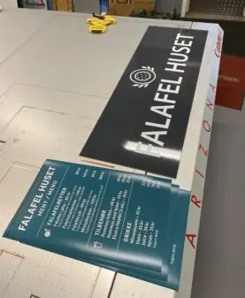 Et stort svart skilt for Falafel Huset og trykte menyer ligger på et bord, der klistremerker er brukt på skiltet.