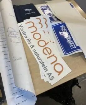 Ulike klistremerker med bedriftslogoer, inkludert Modena og Videoverkstedet, ligger på et bord.