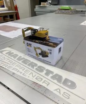 Et stort klistremerke med Haverstad Entreprenør AS logo i sølv ligger på et bord, med en CAT gravemaskinmodell på en eske ved siden av.