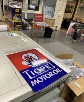 Et stort Tiopet Motor Oil-skilt med rød og blå bakgrunn ligger på en arbeidsbenk i et verksted.