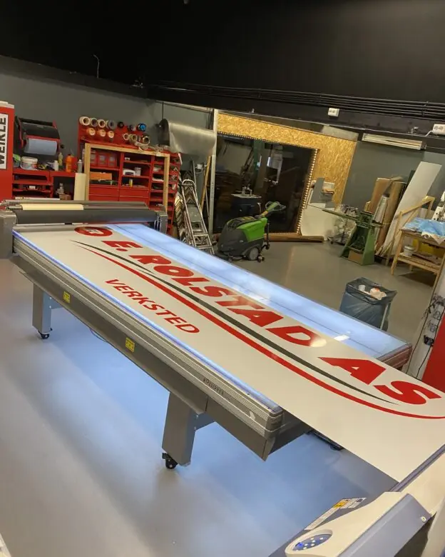 Et stort hvitt ark med røde og svarte klistremerker som staver 'DE ROLSTAD AS VERKSTED' ligger på et opplyst monteringsbord i et verksted.