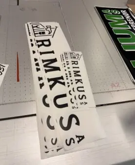 Hvite rektangulære klistremerker med svart Rimkusa-logo og tekst ligger stablet på en arbeidsflate.