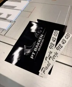 To svarte klistremerker med hvit tekst og logo ligger på en metalloverflate.
