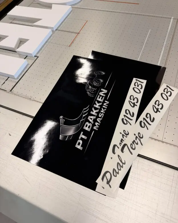 To svarte klistremerker med hvit tekst og logo ligger på en metalloverflate.
