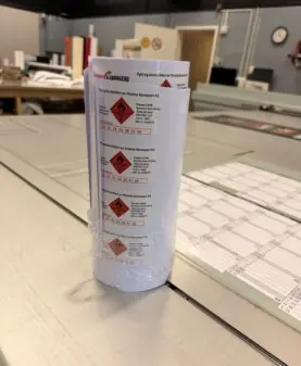 En rull med hvite klistremerker med røde faresymboler ligger på et grått arbeidsbord.