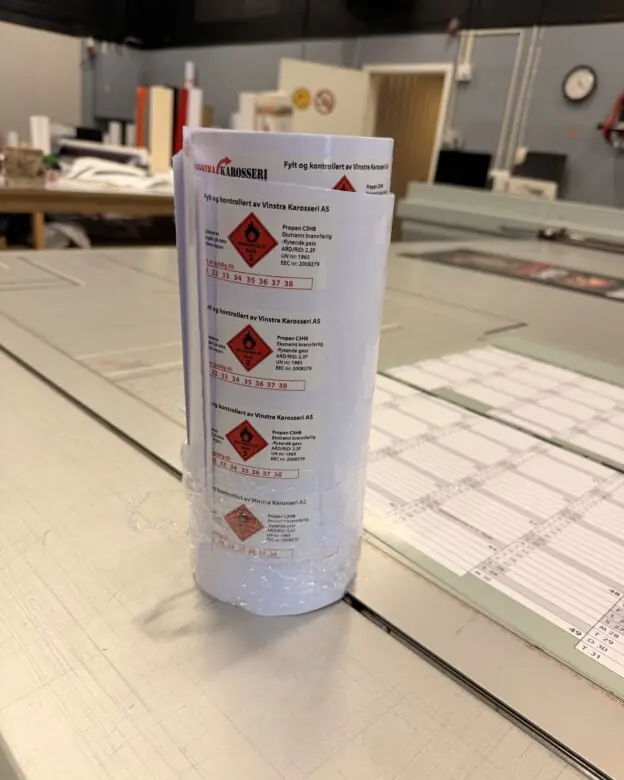 En rull med hvite klistremerker med røde faresymboler ligger på et grått arbeidsbord.