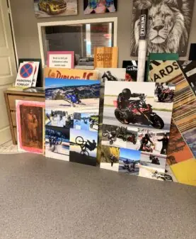 Flere store bilder og plakater med motiver av motorsykler og ekstremsport står lent mot en vegg i et rom.