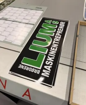 Et rektangulært svart klistremerke med grønn og hvit tekst for Brødrene Lumås Maskinentreprenør ligger på en lys overflate.