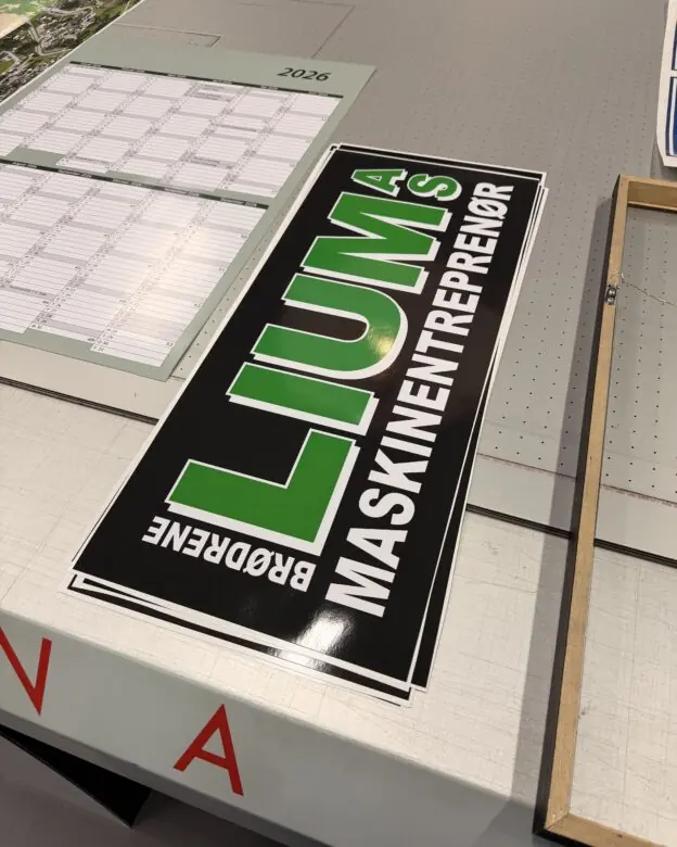 Et rektangulært svart klistremerke med grønn og hvit tekst for Brødrene Lumås Maskinentreprenør ligger på en lys overflate.