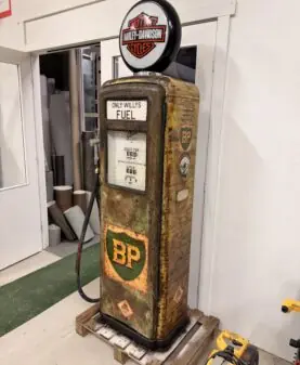 En rusten bensinpumpe med klistremerker og et opplyst Harley-Davidson-skilt på toppen står på en trepall.