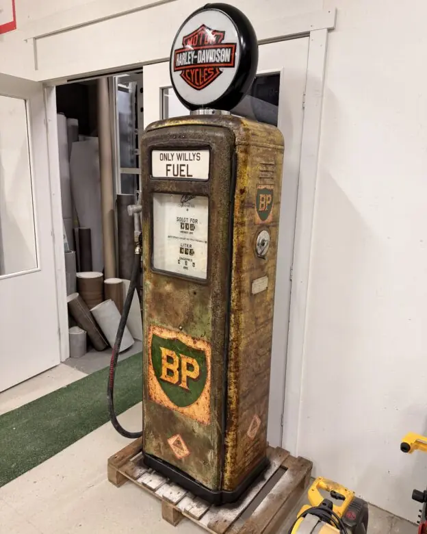 En rusten bensinpumpe med klistremerker og et opplyst Harley-Davidson-skilt på toppen står på en trepall.