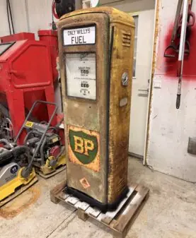 En rusten, gul og grønn bensinpumpe med klistremerker som viser 'BP' og 'ONLY WILLYS FUEL' står på en trepall.