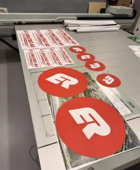 Røde og hvite klistremerker med ER Produkter AS-logo og firmanavn ligger utstilt på et stort bord.
