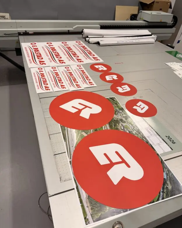 Røde og hvite klistremerker med ER Produkter AS-logo og firmanavn ligger utstilt på et stort bord.