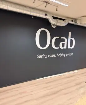 Et stort klistremerke med Ocab-logoen og slagordet "Saving value, helping people" er montert på en mørkegrå vegg i et rom med tregulv.