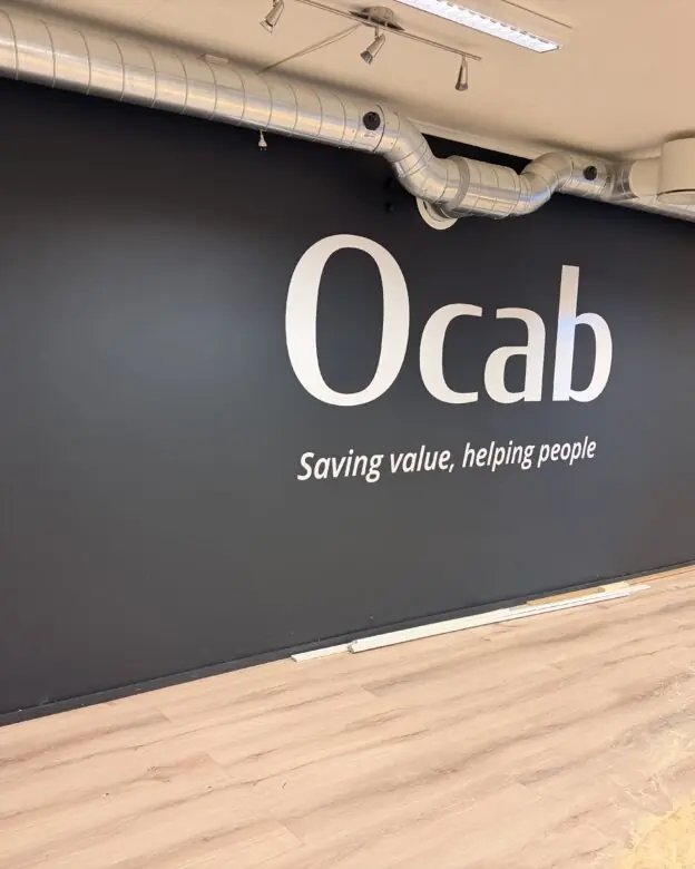 Et stort klistremerke med Ocab-logoen og slagordet "Saving value, helping people" er montert på en mørkegrå vegg i et rom med tregulv.