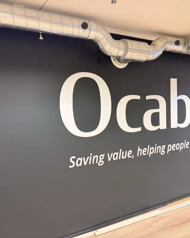 Store hvite klistremerker med Ocab-logo og slagord på en mørkegrå vegg.