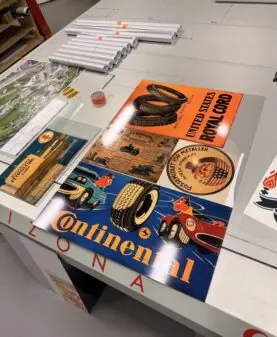 Vintage klistremerker med bil- og dekkreklame ligger utstilt på et arbeidsbord i et trykkeri.