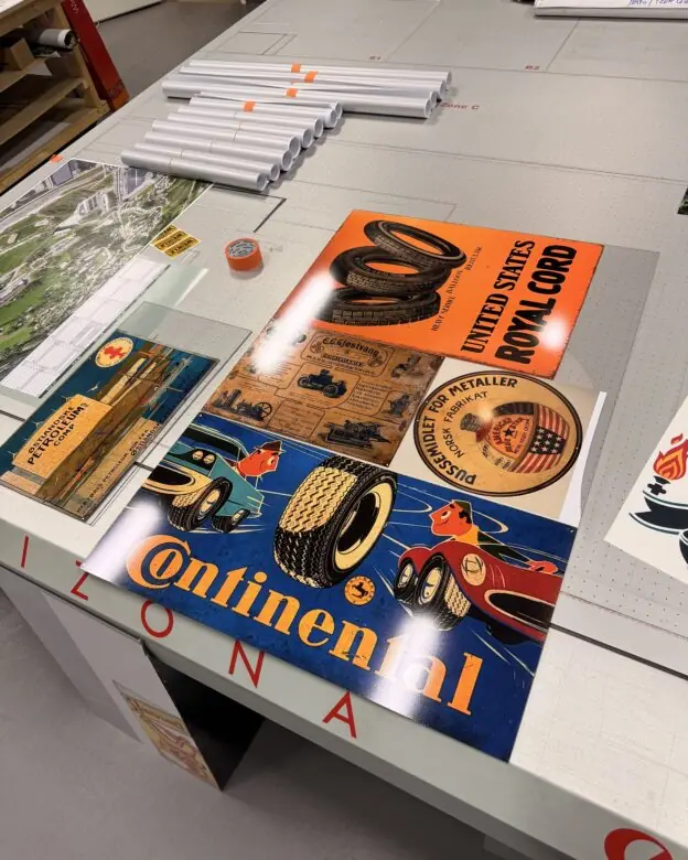 Vintage klistremerker med bil- og dekkreklame ligger utstilt på et arbeidsbord i et trykkeri.