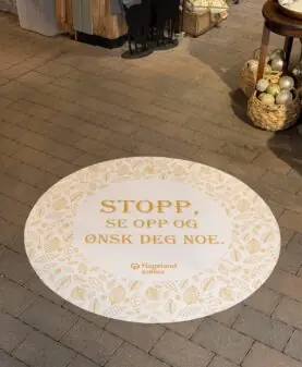 Et rundt gulvklistremerke med teksten "STOPP, SE OPP OG ØNSK DEG NOE." ligger på flisgulvet i en butikk.