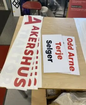 Flere hvite klistremerker med rød tekst som viser "AKERSHUS" og navneskilt som "Odd Arne" og "Terje Selger" ligger på et trebord.