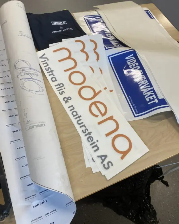Ulike klistremerker med bedriftslogoer, inkludert Modena og Videoverkstedet, ligger på et bord.