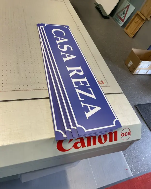 Flere blå klistremerker med hvit tekst "CASA REZA" ligger stablet på en Canon Océ plotter.