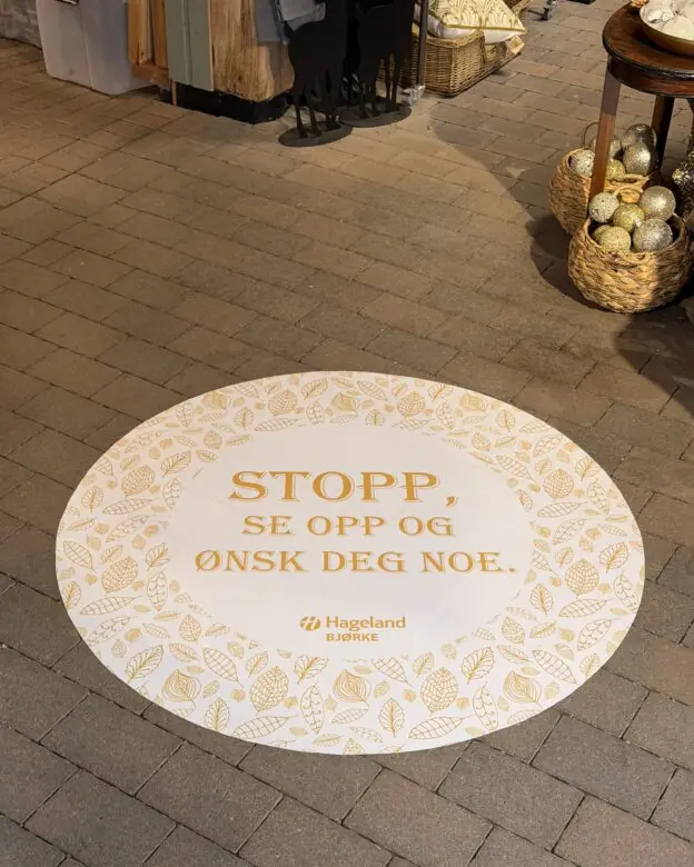 Et rundt gulvklistremerke med teksten "STOPP, SE OPP OG ØNSK DEG NOE." ligger på flisgulvet i en butikk.