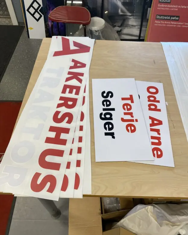 Flere hvite klistremerker med rød tekst som viser "AKERSHUS" og navneskilt som "Odd Arne" og "Terje Selger" ligger på et trebord.