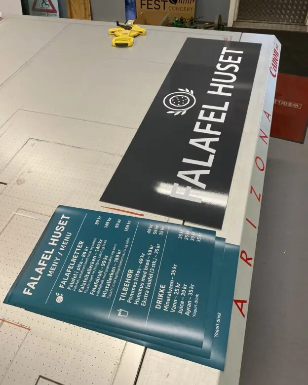 Et stort svart skilt for Falafel Huset og trykte menyer ligger på et bord, der klistremerker er brukt på skiltet.
