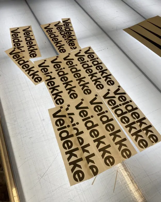 Brune klistremerker med svart Veidekke-logo ligger på et opplyst bord.