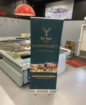 En mørkegrønn rollup med logoen Per Gynt Hjortekjøtt og illustrasjon av hjort står på et verkstedgulv.