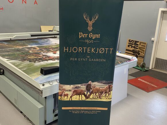 En mørkegrønn rollup med logoen Per Gynt Hjortekjøtt og illustrasjon av hjort står på et verkstedgulv.