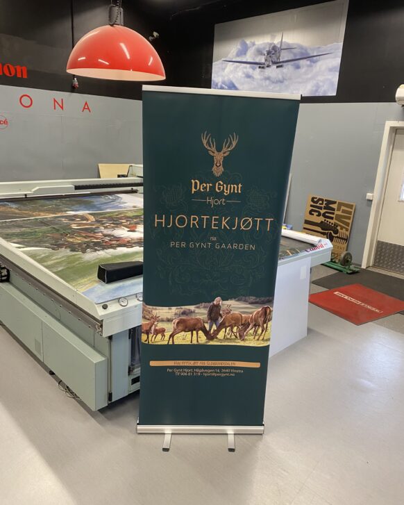 En mørkegrønn rollup med logoen Per Gynt Hjortekjøtt og illustrasjon av hjort står på et verkstedgulv.