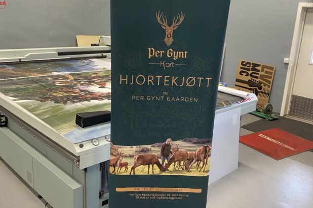 En mørkegrønn rollup med logoen Per Gynt Hjortekjøtt og illustrasjon av hjort står på et verkstedgulv.