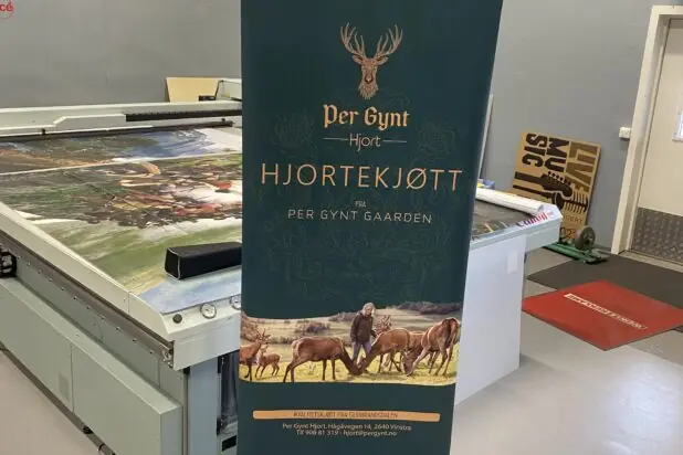 En mørkegrønn rollup med logoen Per Gynt Hjortekjøtt og illustrasjon av hjort står på et verkstedgulv.