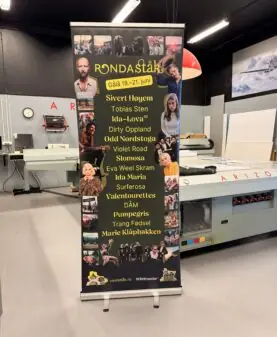 En svart rollup-banner for Rondaståkk festival med artistnavn og små bilder står i et trykkeri.