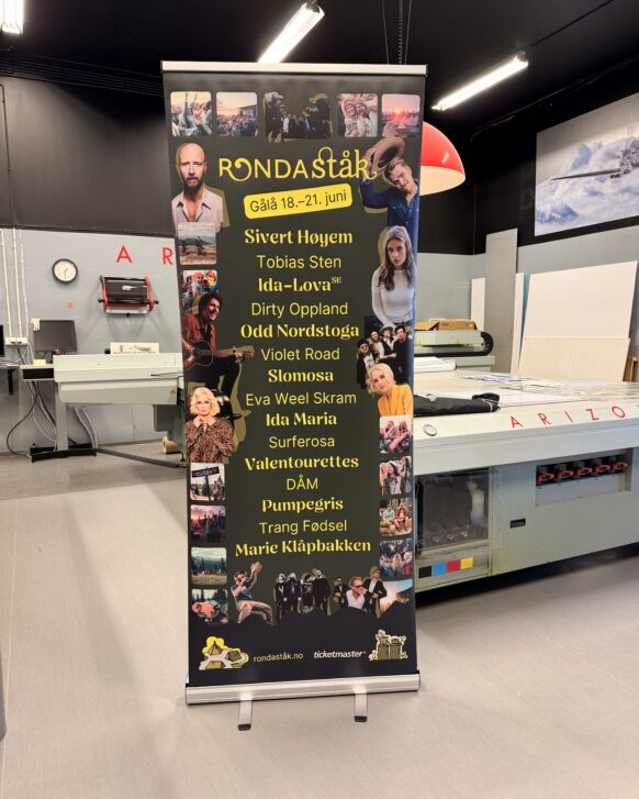 En svart rollup-banner for Rondaståkk festival med artistnavn og små bilder står i et trykkeri.