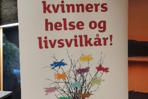 En hvit rollup med rød tekst som oppfordrer til å støtte Sanitetens arbeid for kvinners helse og livsvilkår.