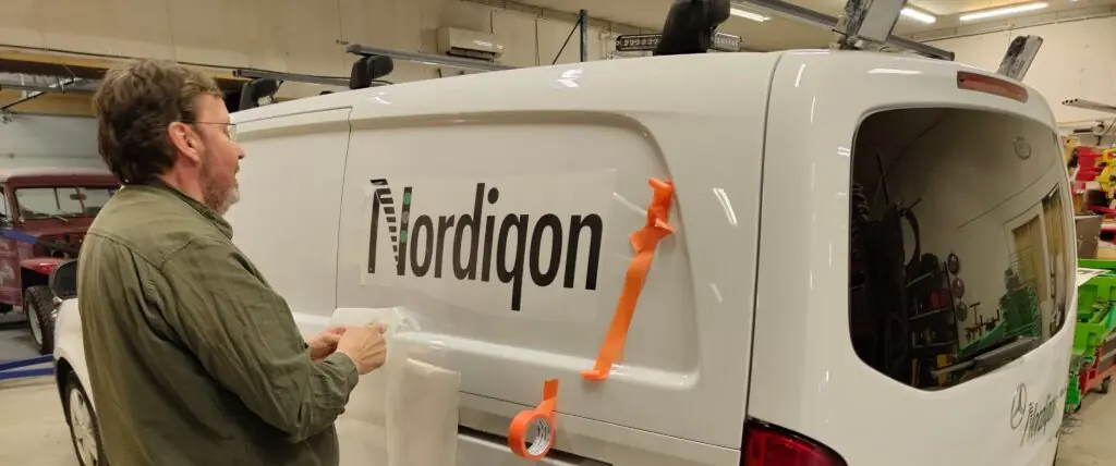 En mann med skjegg og grønn skjorte folierer siden på en hvit varebil med Nordiqon-logoen i et verksted.