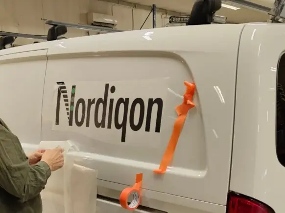 En mann med skjegg og grønn skjorte folierer siden på en hvit varebil med Nordiqon-logoen i et verksted.