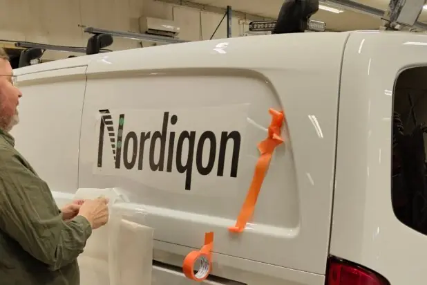 En mann med skjegg og grønn skjorte folierer siden på en hvit varebil med Nordiqon-logoen i et verksted.